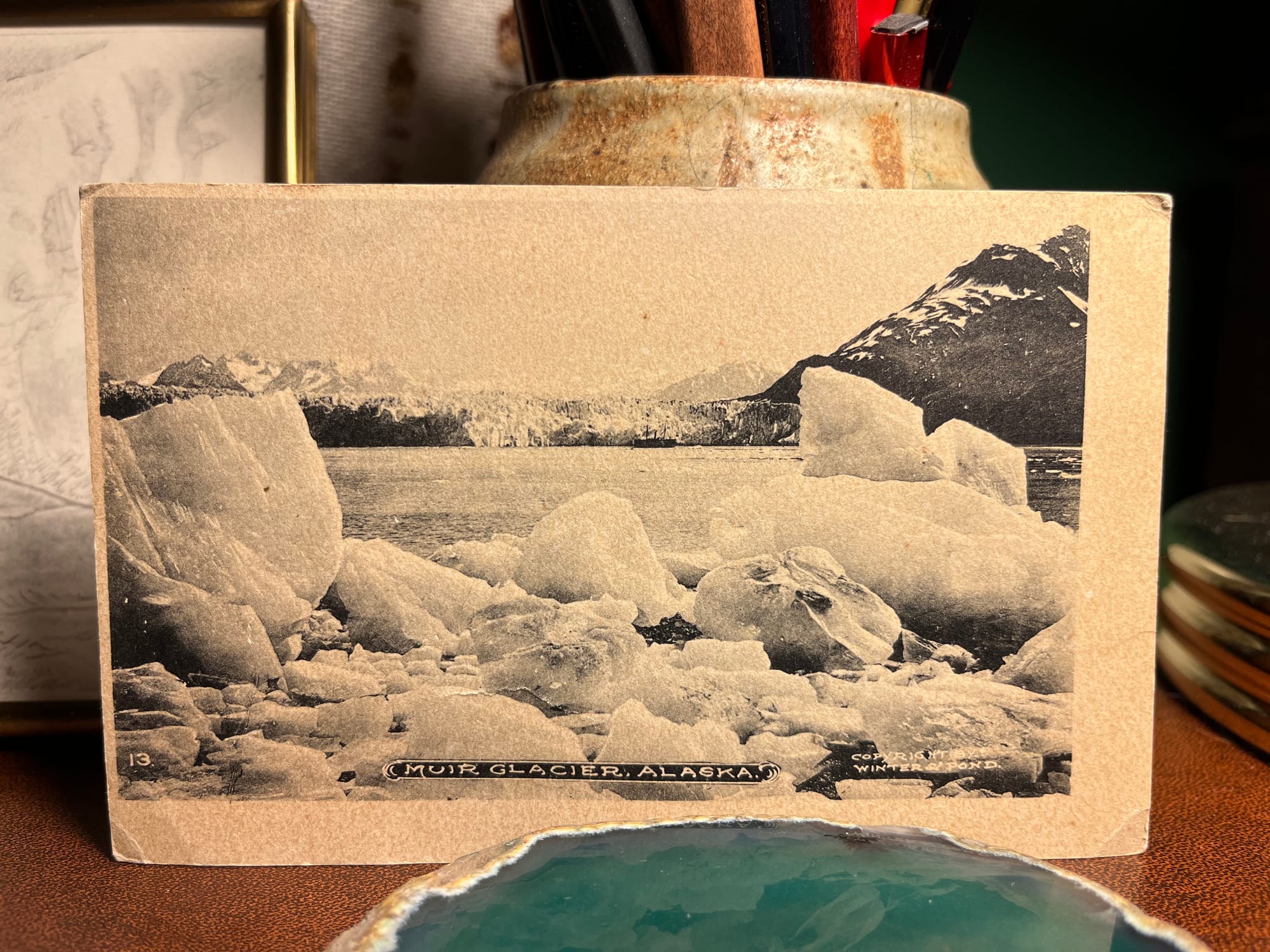 A vintage, black & white postcard with the caption “Muir Glacier, Alaska” sits propped against a ceramic pencil holder on a desk.
